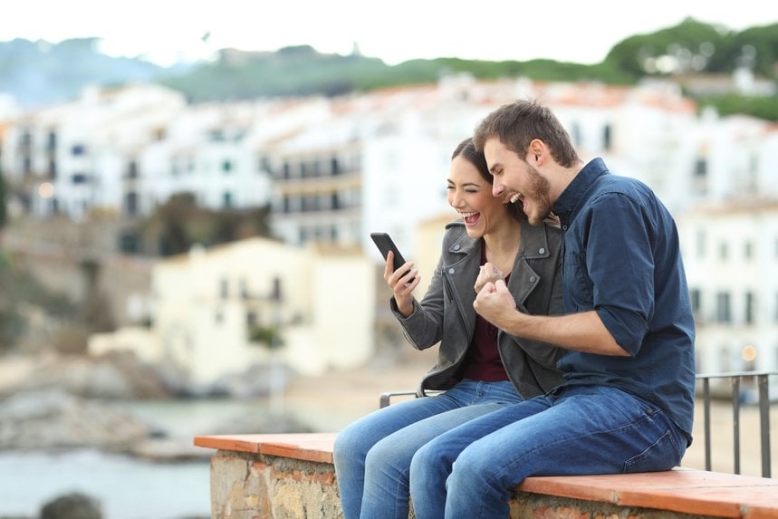Two travellers excitedly looking at a phone on a terrace in a whitewashed seaside town.