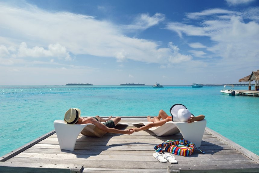 Two people sunbathing on loungers on a jetty over turquoise water.