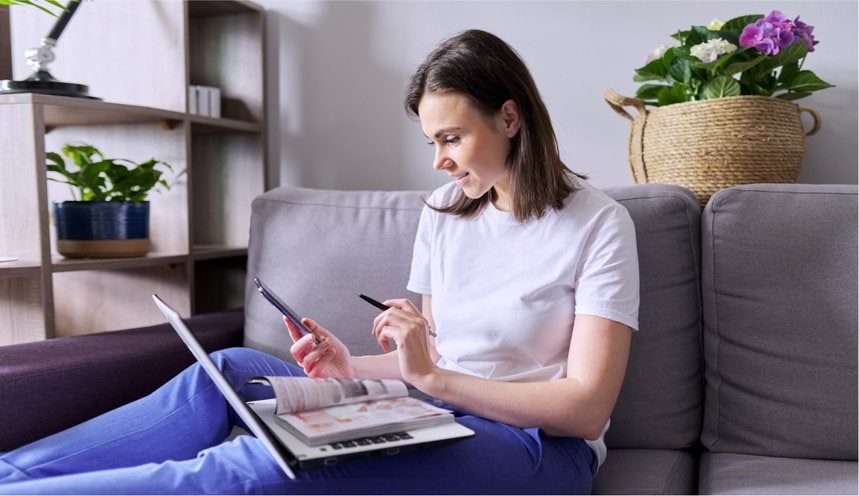 A woman on a sofa using her phone, with a magazine open on her laptop.